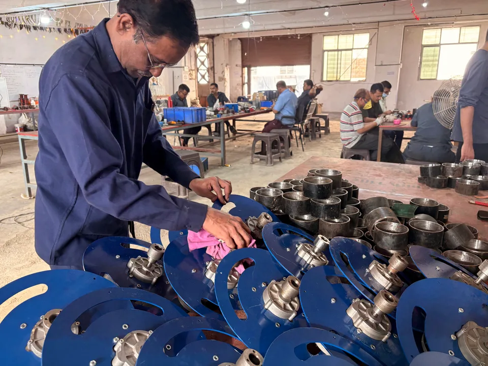 Operator working at a bench with metal and blue-painted assemblies while other workers sit at nearby benches